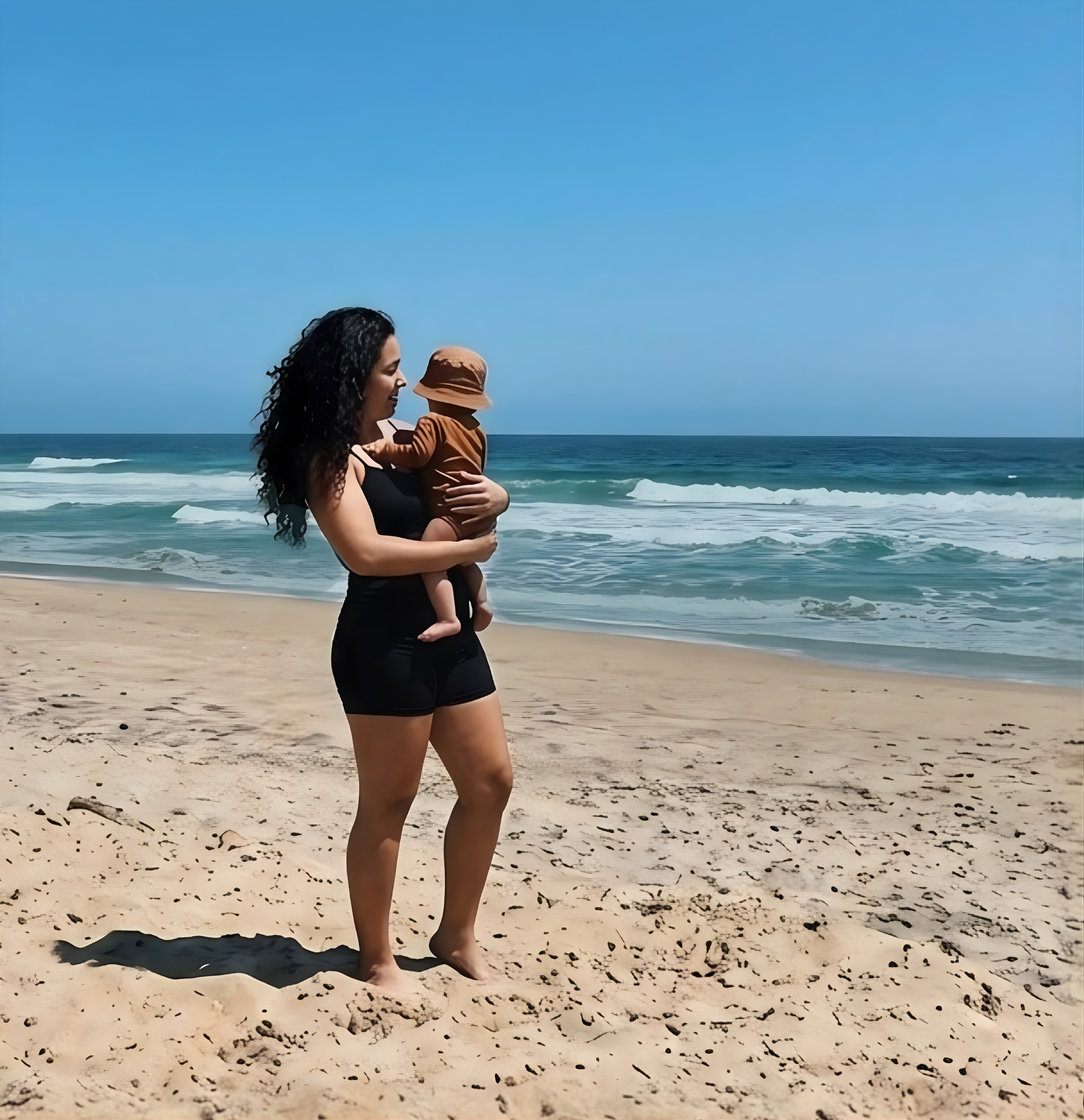 Une maman épanouie sur la plage tenant son bébé dans ses bras. Elle porte un ensemble de bain semi-couvrant composé d'un short et d'un top de la marque Eclypse, profitant pleinement d'un moment de promenade avec son enfant en toute sérénité